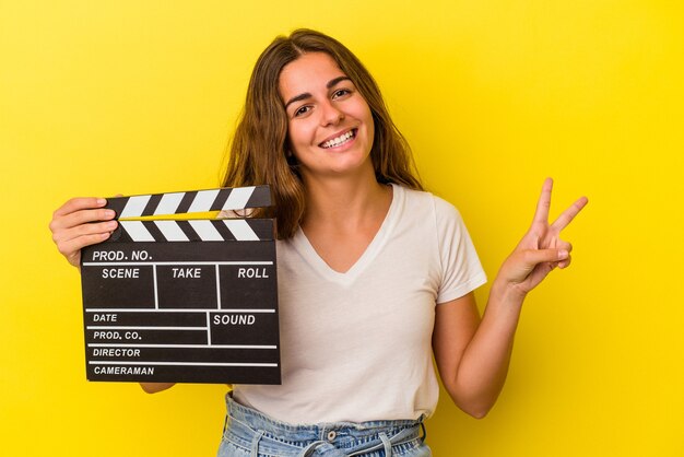 Jeune femme caucasienne tenant un clap isolé sur fond jaune joyeux et insouciant montrant un symbole de paix avec les doigts.