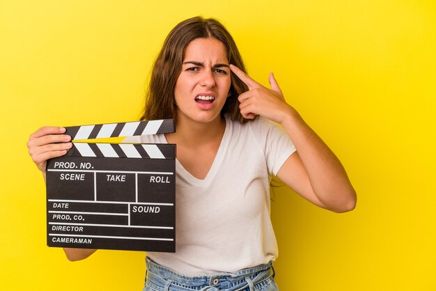 Jeune femme caucasienne tenant un clap isolé sur fond jaune montrant un geste de déception avec l'index.