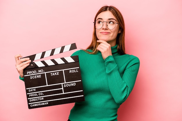 Jeune femme caucasienne tenant un clap isolé sur fond rose regardant de côté avec une expression douteuse et sceptique