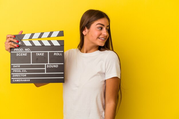 Jeune femme caucasienne tenant un clap isolé sur un mur blanc regarde de côté souriante, gaie et agréable.