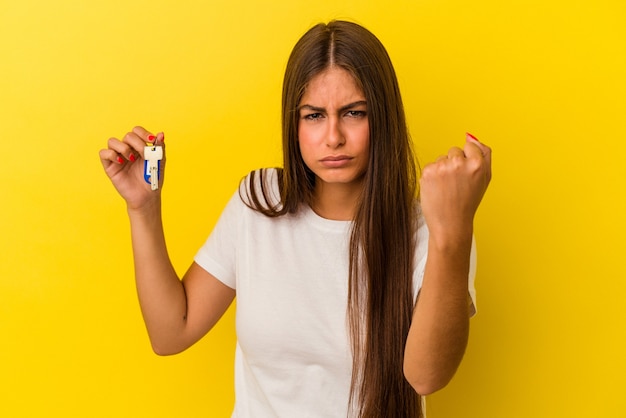 Jeune femme caucasienne tenant une clé d'accueil isolée sur fond jaune montrant le poing à la caméra, expression faciale agressive.