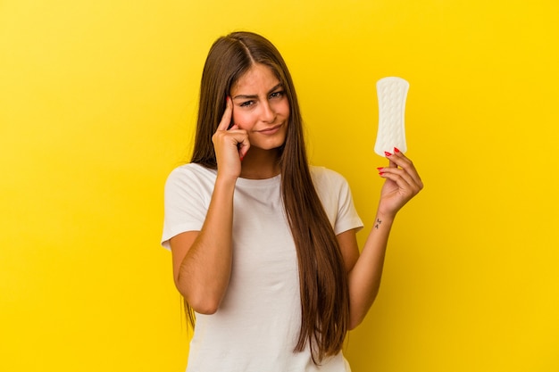 Jeune femme caucasienne tenant une compresse isolée sur fond jaune pointant le temple avec le doigt, pensant, concentrée sur une tâche.