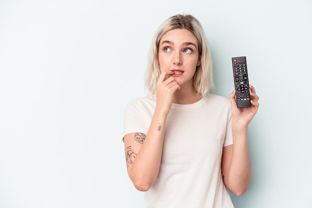 Jeune femme caucasienne tenant un contrôleur de télévision isolé sur fond bleu détendue en pensant à quelque chose en regardant un espace de copie.