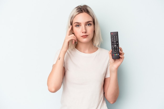 Jeune femme caucasienne tenant un contrôleur de télévision isolé sur fond bleu pointant le temple avec le doigt, pensant, concentré sur une tâche.