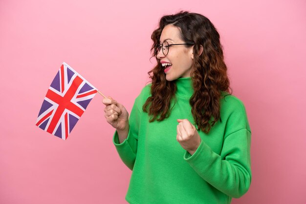 Jeune femme caucasienne tenant un drapeau anglais isolé sur fond rose célébrant une victoire