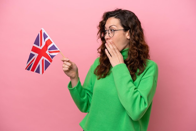 Jeune femme caucasienne tenant un drapeau anglais isolé sur fond rose avec une expression faciale surprise et choquée