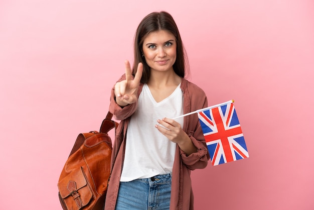 Jeune femme caucasienne tenant un drapeau du Royaume-Uni isolé sur fond rose souriant et montrant le signe de la victoire