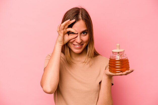 Jeune femme caucasienne tenant du miel isolé sur fond rose excité en gardant le geste ok sur les yeux