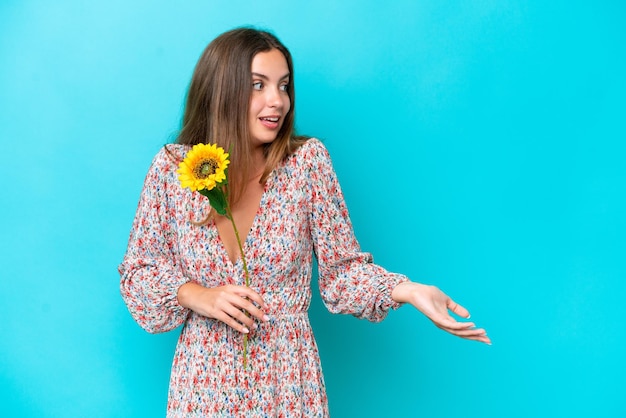 Jeune femme caucasienne tenant du tournesol isolé sur fond bleu avec une expression surprise tout en regardant de côté