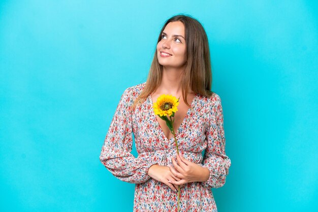 Jeune femme caucasienne tenant du tournesol isolé sur fond bleu pensant à une idée tout en levant les yeux