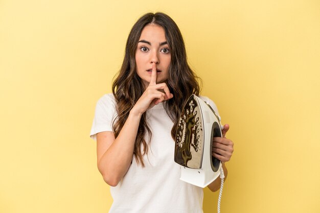 Jeune femme caucasienne tenant un fer isolé sur fond jaune gardant un secret ou demandant le silence.