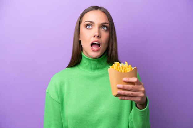 Jeune femme caucasienne tenant des frites frites sur fond violet levant les yeux et avec une expression surprise