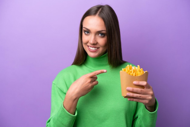 Jeune femme caucasienne tenant des frites frites sur fond violet pointant vers le côté pour présenter un produit