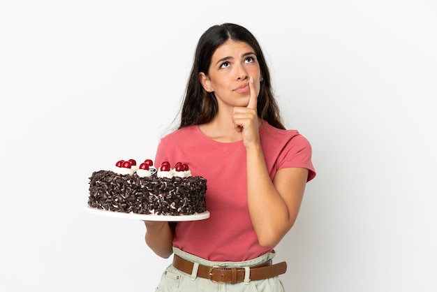 Jeune femme caucasienne tenant un gâteau d'anniversaire isolé sur fond blanc ayant des doutes en levant les yeux