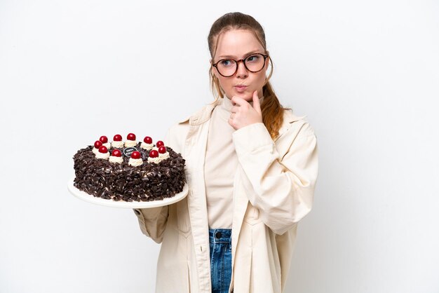 Jeune femme caucasienne tenant un gâteau d'anniversaire isolé sur fond blanc ayant des doutes et pensant