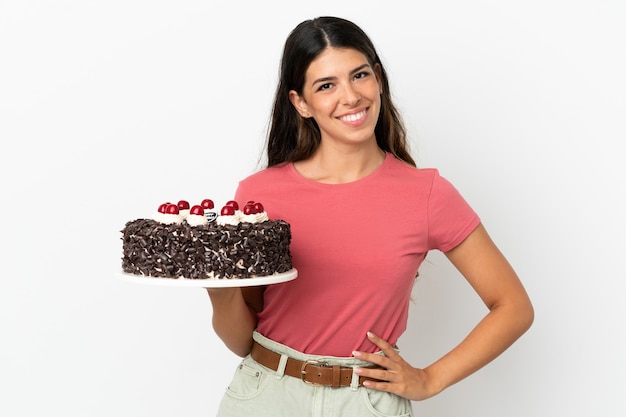 Jeune femme caucasienne tenant un gâteau d'anniversaire isolé sur fond blanc posant avec les bras à la hanche et souriant