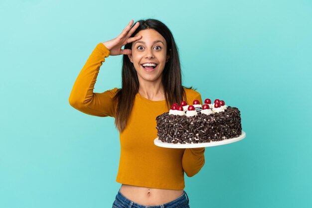 Jeune femme caucasienne tenant un gâteau d'anniversaire isolé sur fond bleu avec une expression de surprise