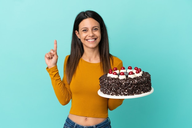 Jeune femme caucasienne tenant un gâteau d'anniversaire isolé sur fond bleu montrant et levant un doigt en signe du meilleur