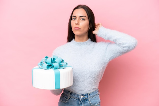 Jeune femme caucasienne tenant un gâteau d'anniversaire isolé sur fond rose ayant des doutes