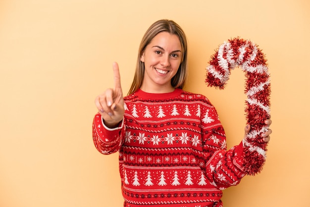 Jeune femme caucasienne tenant un gros bâton de Noël isolé sur fond jaune montrant le numéro un avec le doigt.