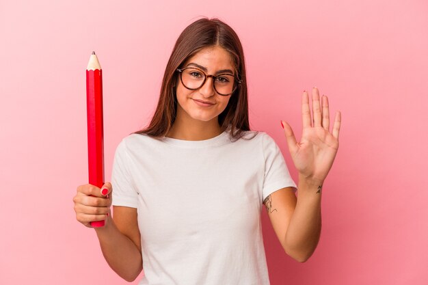 Jeune femme caucasienne tenant un gros crayon isolé sur fond rose souriant joyeux montrant le numéro cinq avec les doigts.