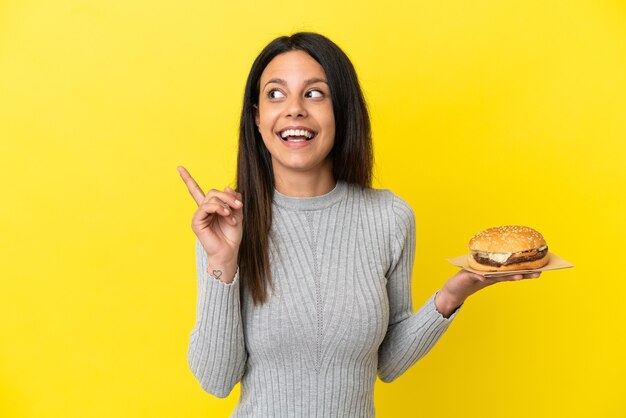 Jeune femme caucasienne tenant un hamburger isolé sur fond jaune dans l'intention de réaliser la solution tout en levant un doigt vers le haut