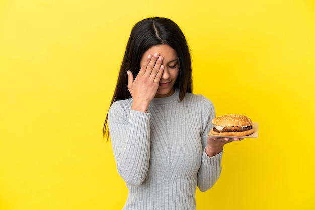 Jeune femme caucasienne tenant un hamburger isolé sur fond jaune avec une expression fatiguée et malade