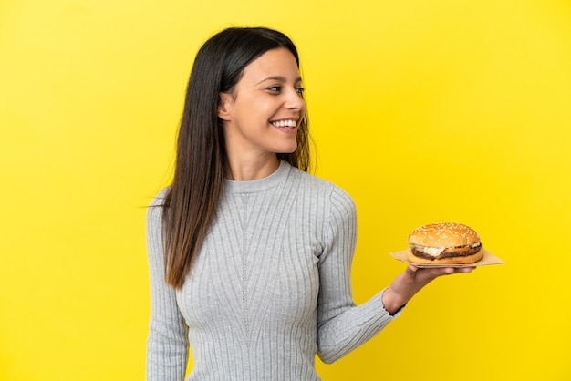 Jeune femme caucasienne tenant un hamburger isolé sur fond jaune regardant sur le côté et souriant