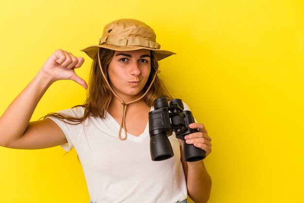 Jeune femme caucasienne tenant des jumelles isolées sur fond jaune montrant un geste d'aversion, les pouces vers le bas. Notion de désaccord.