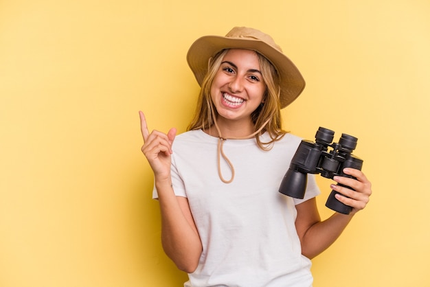 Jeune femme caucasienne tenant des jumelles isolées sur fond jaune souriant et pointant de côté, montrant quelque chose dans un espace vide.