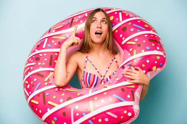 Jeune femme caucasienne tenant un matelas pneumatique isolé sur fond bleu pointant vers le haut avec la bouche ouverte