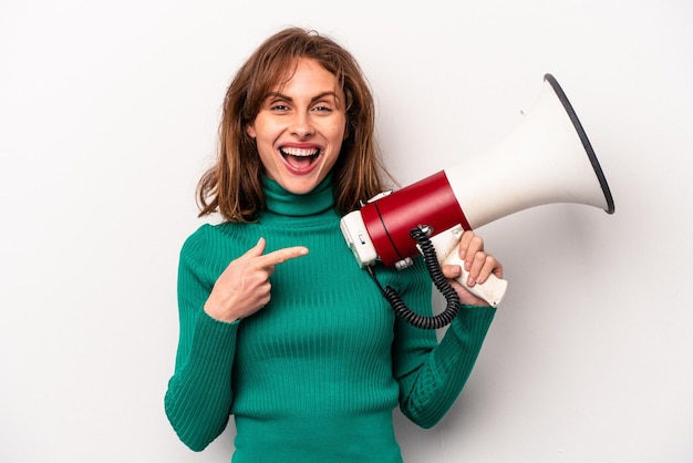 Jeune femme caucasienne tenant un mégaphone isolé sur fond blanc souriant et pointant de côté montrant quelque chose à l'espace vide