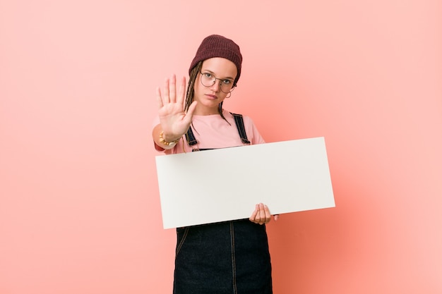 Jeune femme caucasienne tenant une pancarte debout avec la main tendue montrant le panneau d'arrêt, vous empêchant.