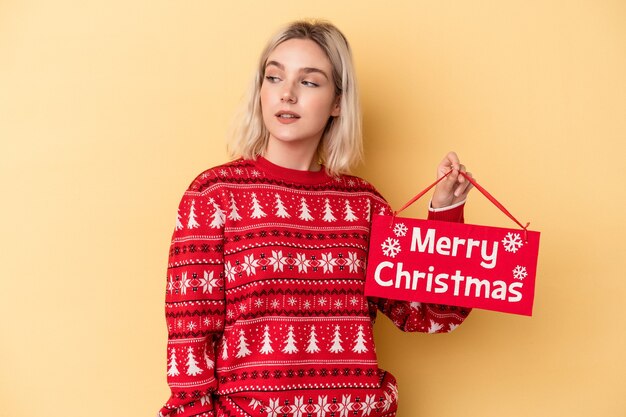 Jeune femme caucasienne tenant une pancarte de joyeux noël isolée sur fond jaune regarde de côté souriante, gaie et agréable.