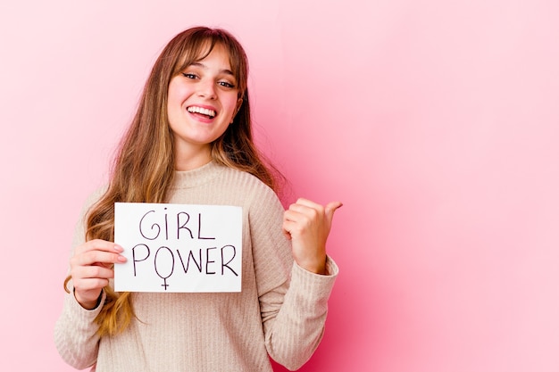 Jeune femme caucasienne tenant une pancarte de puissance de fille isolée sur les points de mur rose avec le pouce, en riant et sans soucis.