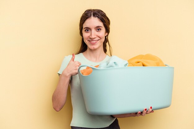 Jeune femme caucasienne tenant un panier à linge isolé sur fond jaune souriant et levant le pouce vers le haut