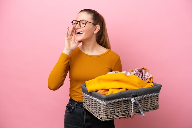 Jeune femme caucasienne tenant un panier à linge isolé sur fond rose criant avec la bouche grande ouverte sur le côté