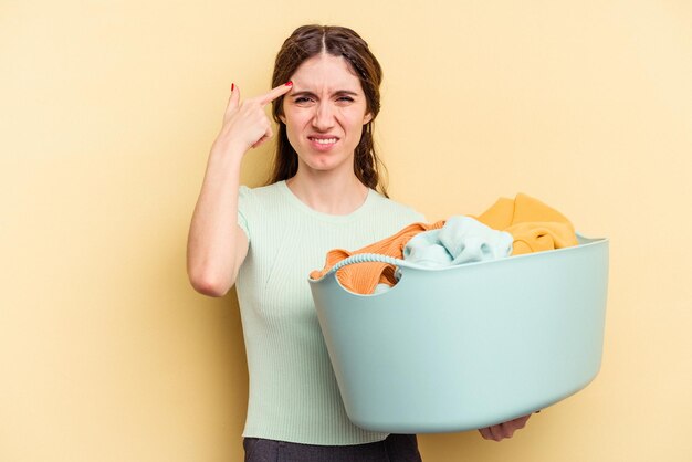 Jeune femme caucasienne tenant un panier à vêtements isolé sur fond jaune montrant un geste de déception avec l'index