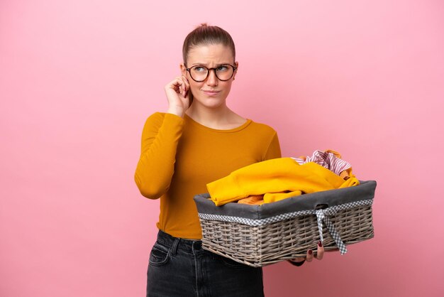 Jeune femme caucasienne tenant un panier à vêtements isolé sur fond rose frustré et couvrant les oreilles