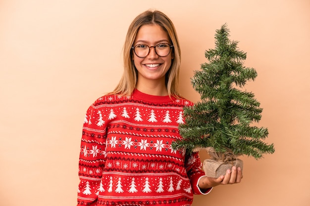 Jeune femme caucasienne tenant un petit arbre de Noël isolé sur fond beige heureux, souriant et joyeux.