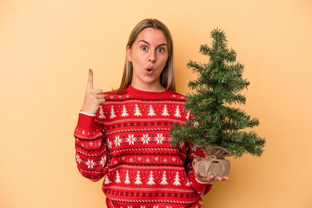 Jeune femme caucasienne tenant un petit arbre de noël isolé sur fond jaune ayant une bonne idée, concept de créativité.