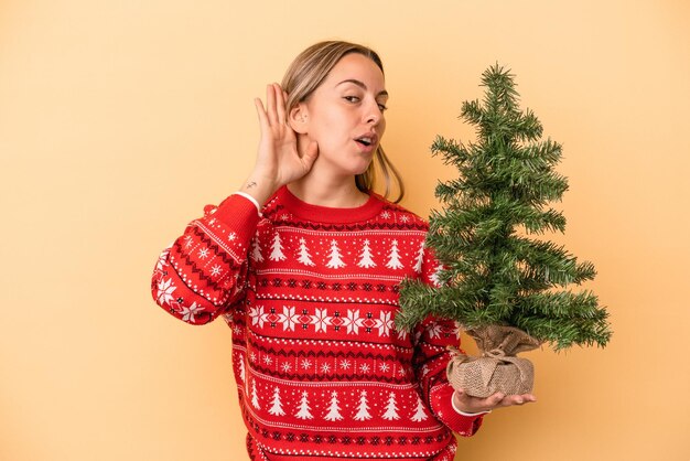 Jeune femme caucasienne tenant un petit arbre de Noël isolé sur fond jaune essayant d'écouter un potin.