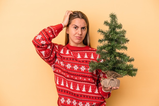 Jeune femme caucasienne tenant un petit arbre de noël isolé sur fond jaune étant choquée, elle s'est souvenue d'une réunion importante.