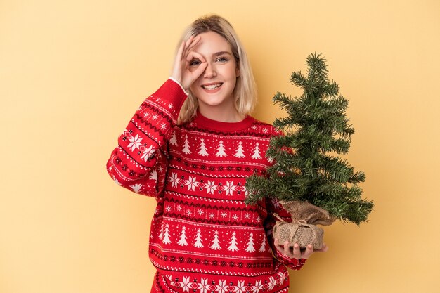 Jeune femme caucasienne tenant un petit arbre de Noël isolé sur fond jaune excité en gardant le geste ok sur les yeux.