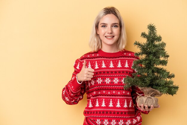 Jeune femme caucasienne tenant un petit arbre de Noël isolé sur fond jaune souriant et levant le pouce vers le haut