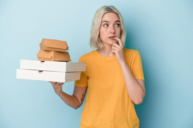 Jeune femme caucasienne tenant des pizzas et des hamburgers isolés sur fond bleu détendue en pensant à quelque chose en regardant un espace de copie.