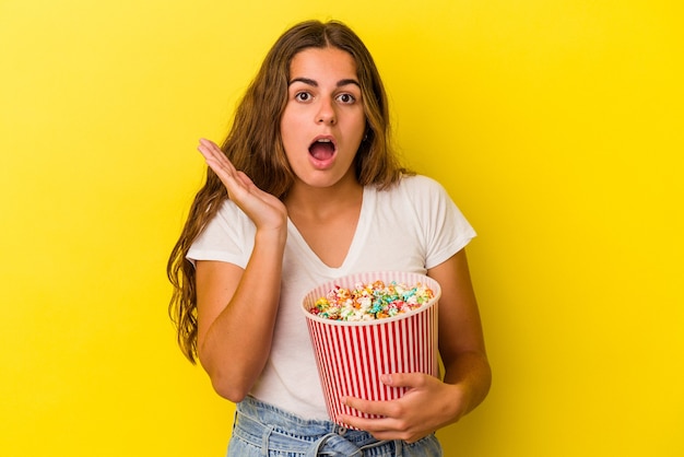 Jeune femme caucasienne tenant un pop-corn isolé sur fond jaune surpris et choqué.