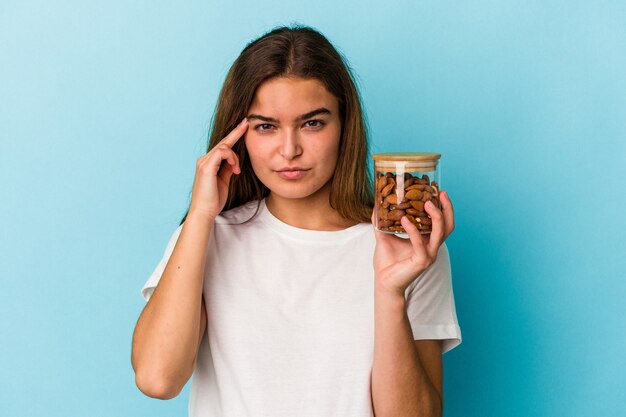 Jeune femme caucasienne tenant un pot d'amande isolé sur fond bleu pointant le temple avec le doigt, pensant, concentré sur une tâche.