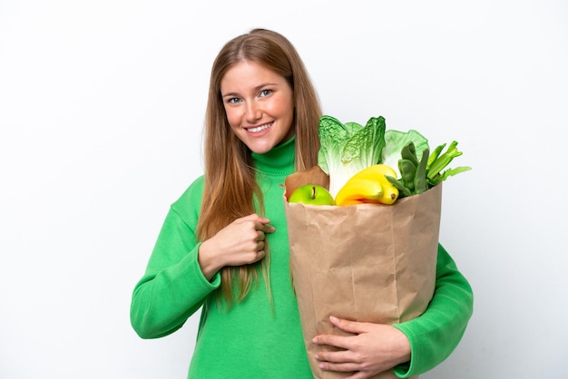 Jeune femme caucasienne tenant un sac d'épicerie isolé sur fond blanc avec une expression faciale surprise