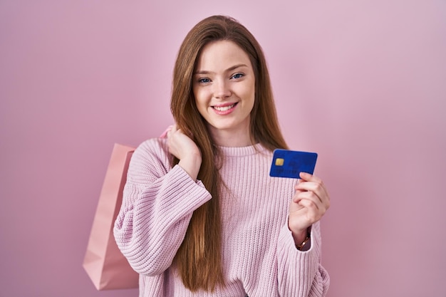 Jeune femme caucasienne tenant un sac à provisions et une carte de crédit souriant avec un sourire heureux et cool sur le visage. montrant les dents.